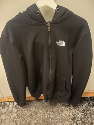 Svart kofta med dragkedja North Face - Säljer en svart kofta från The North Face med dragkedja framtill och vit logga på bröstet. Koftan har huva med snörning och två stora fickor framtill. Perfekt för en avslappnad och sportig stil. Koftan passar även om man har storlek L.