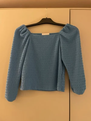 Blå smockad topp från H&M - Supersöt blå topp från H&M med smockad struktur och långa puffärmar. Toppen har en fyrkantig ringning och är perfekt för dig som gillar trendiga detaljer och en bekväm passform.