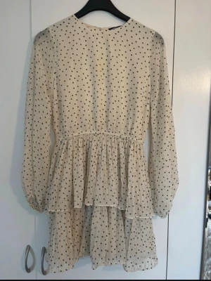 Prickig volangklänning i beige - Supersöt kortklänning i beige med svarta prickar och volanger. Klänningen har långärmade ärmar och är tillverkad i ett lätt, transparent material som ger en luftig känsla. Perfekt för dig som gillar romantisk och lekfull stil.