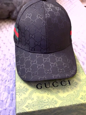 Svart Gucci keps med GG-mönster - Snygg svart keps från Gucci med klassiskt GG-monogram över hela kepsen och gröna/röda ränder på sidan. Justerbar baktill och insida med Gucci-logga. Tillverkad i slitstarkt tyg som ger en exklusiv känsla.