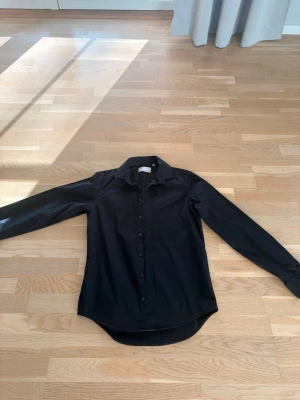 Svart skjorta från Casual Friday - Klassisk svart skjorta från Casual Friday med lång ärm och knappar framtill. Skjortan har en stilren krage och är tillverkad i ett mjukt bomullstyg som känns bekvämt mot huden. Perfekt för dig som gillar en enkel och clean look.