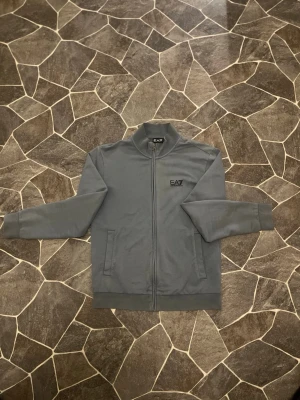 EA7 Armani Zip Ljusblå - En mycket fin vän använd zip ifrån EA7 Armani. Tröjan har använts ett par gånger men är generellt i fint skick. På sista bilden ser man ett märke där back, kanske går och tvätta bort.   Priset kan diskuteras!