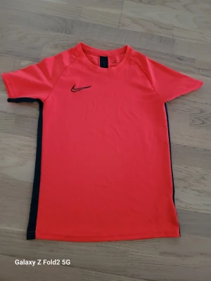 Röd Nike Dri-Fit t-shirt - Snygg röd Nike Dri-Fit t-shirt med svart logga på bröstet och svarta detaljer längs sidorna. Tillverkad i ett lätt och funktionellt material som passar perfekt för träning. Klassisk passform och korta ärmar.