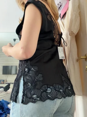 Svart spetstopp - Supersnygg svart linnetopp med djup v-ringning och spetsdetaljer längs kanterna. Toppen har snörning i ryggen. Aldrig använd med prislapp. Storlek M men passar S.💕