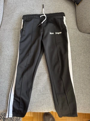 Svarta mjukisbyxor Palm Angels - Säljer ett par svarta mjukisbyxor från Palm Angels med vita sidoränder och snörning i midjan. Byxorna har två fickor och logga tryckt på vänster sida. Perfekta för en chill och sportig look.