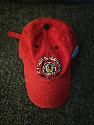 Röd Chicago Blackhawks keps Adidas - Snygg röd keps med Chicago Blackhawks-logga framtill och broderad text. Justerbar rem baktill och Adidas-märke på sidan. Perfekt för hockeyfans som vill sticka ut med stilren accessoar.