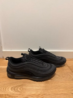 Nike Air Max 97 helsvarta sneakers - Säljer ett par helsvarta Nike Air Max 97 sneakers med den ikoniska vågiga designen och synlig Air-enhet i sulan. Skorna har en låg silhuett, dragflikar på plös och häl samt ovandel i mesh och syntetmaterial. Perfekta för dig som gillar stilrena och sportiga sneakers.