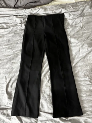 Svarta utsvängda kostymbyxor - Säljer ett par svarta kostymbyxor från Gina tricot med utsvängda ben och pressveck. Byxorna har en stilren, klassisk look som passar perfekt till en snygg outfit. Materialet känns mjukt och har en lätt stretch för extra komfort.