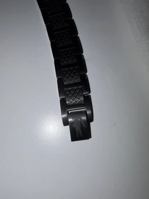 Svart länkarmband från Lacoste - Snyggt svart länkarmband till klocka från Lacoste med graverad krokodil på spännet och diskret mönster på länkarna. Armbandet är tillverkat i metall och levereras med originalask och tygpåse med Lacoste-logga.