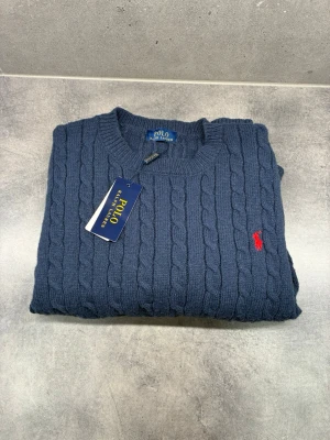 Stickad tröja från polo  - Kabelstickad mörkblå tröja från Polo Ralph Lauren med rund halsringning och röd broderad logga på bröstet. Tröjan har ribbade muddar vid ärmslut och nederkant. Klassisk och tidlös design i mjukt material.