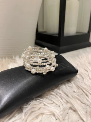 Silvrigt armband med pärlor - Snyggt armband i spiralform med vita pärlor och små glittrande stenar på silverfärgad metalltråd. Armbandet har flera lager och ger en elegant look med sina olika storlekar på pärlorna. Perfekt accessoar för att lyfta din outfit.