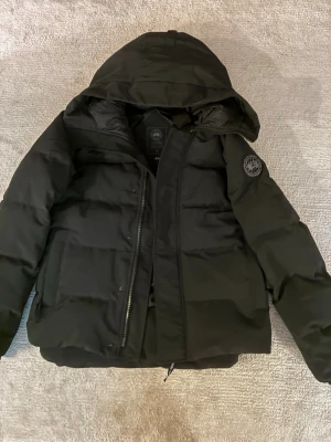 Svart dunjacka från Canada Goose - Säljer en svart dunjacka från Canada Goose med huva och diskret logotyp på ärmen. Jackan har en rak passform, dragkedja framtill och stora fickor. Perfekt för kalla vinterdagar och riktigt snygg till streetwear-stilen.