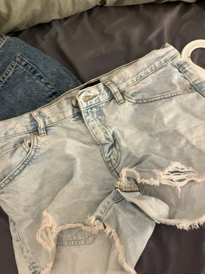 Ljusblå slitna jeansshorts - Säljer ett par ljusblå jeansshorts med råa, slitna detaljer och fransar vid bensluten. Shortsen har klassisk femficksmodell, bälteshällor och knappgylf. Perfekta för en avslappnad och trendig look.
