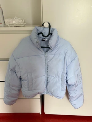 Ljusblå cropped pufferjacka - Säljer en ljusblå cropped pufferjacka med hög krage och dragkedja framtill. Jackan har två fickor på sidorna och breda, quiltade paneler för extra volym. Perfekt för kalla vinterdagar och ger en trendig look.