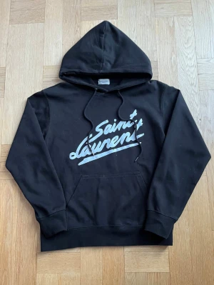 Svart Saint Laurent hoodie - Snygg svart hoodie från Saint Laurent (YSL) i storlek S med stor vit logga framtill. Klassisk modell med huva, dragsnören och stor magficka. Tillverkad i mjukt och bekvämt material som känns skönt mot huden. Hoodien är i mycket bra skick, utan hål, fläckar eller andra defekter. Äkthetsbevis finns. Perfekt för en avslappnad men stilren och trendig look. Hör gärna av dig vid frågor Mvh Olof!