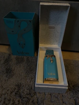 Vulcan parfym från French Avenue - Snygg parfymflaska i turkost med gulddetaljer och ormdekor från French Avenue. Flaskan har en unik, fyrkantig form och levereras i en matchande lyxig presentförpackning med mönstrad insida. Perfekt för dig som gillar exklusiva och iögonfallande dofter.