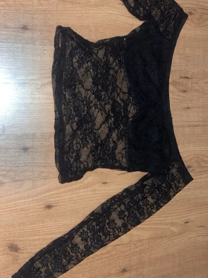 Svart spetsig offshoulder topp - Snygg svart offshoulder topp i genombruten spets med långa ärmar. Toppen är figurnära och har en transparent look som ger en edgy vibe. Perfekt att styla med höga jeans eller kjol för en trendig outfit.