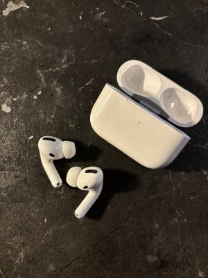 Apple AirPods Pro med laddningsetui - Säljer ett par vita Apple AirPods Pro med trådlöst laddningsetui. Hörlurarna har silikonproppar för bra passform och brusreducering. Allt originaltillbehör som extra öronproppar, laddkabel och förpackning ingår. Snygg och stilren design i blank plast.