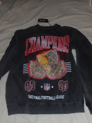 Svart NFL sweatshirt från Boohoo - Cool svart sweatshirt från Boohoo med NFL-tema och stor CHAMPIONS-print i rött och vitt framtill. Tröjan har rund halsringning och tryck med ost och sportmotiv. Perfekt för dig som gillar streetwear och amerikansk fotboll.