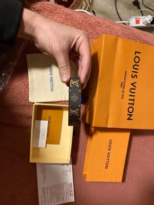 Louis Vuitton monogram armband - Snyggt armband från Louis Vuitton i klassiskt brunt monogrammönster med guldiga LV-detaljer. Tillverkat i läder med ikoniska loggor och stilren design. Perfekt accessoar för dig som vill ha något lyxigt och trendigt från ett av världens mest kända modehus.