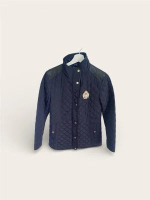 Ralph Lauren Quiltad Jacka – Marinblå med Skinndetaljer (Nyskick!) - Säljer en klassisk och elegant vårjacka från Lauren Ralph Lauren. Modellen är en quiltad "Riding Jacket" i mörkblått (Navy) med exklusiva detaljer. Jackan har snygga förstärkningar i skinnimitation på axlarna och ett broderat emblem med guldtråd på bröstet.  Skick: Nyskick! Jackan är endast provad inomhus, aldrig använd ute. Helt fri från slitage.  Storlek: Small (S). (Passform: Figursydd och smickrande). Nypris ca 3 000 kr.