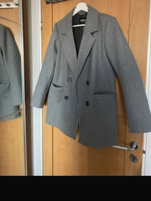 Grå oversized kavaj kappa från ONLY - Stilren grå kavaj kappa från ONLY i oversized modell med dubbelknäppning och breda slag. Kappan har två framfickor och är fodrad. Perfekt för en trendig och avslappnad look. Materialet känns mjukt och har en ull-liknande struktur.