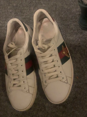 Gucci Ace vita sneakers med bi - Säljer ett par vita Gucci Ace sneakers med klassiska gröna och röda ränder på sidan och en guldbroderad bi. Baksidan har metallicdetaljer i rött och grönt ormskinnsmönster. Skorna har lågt skaft och vita snören. Perfekta för dig som vill sticka ut.