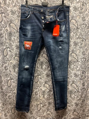 Blå slitna jeans från Dsquared2 - Säljer ett par blå skinny jeans från Dsquared2 med slitningar, patch på vänster lår och knappar i gylfen. Jeansen har en tvättad look, orange sömmar och detaljer som ger en cool streetkänsla. Perfekta för dig som gillar en trendig och avslappnad stil.