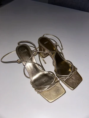 Guldiga klackar från Mango - Super snygga guldiga klackar från Mango (MNG)! Storlek 38. Nypris 399kr. En tjock klack så de är jätte sköna att gå i. Använda fåtal gånger. Långa band som går att snurra runt foten/benet med spännen.