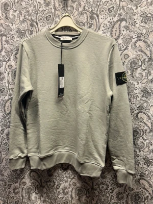 Grå sweatshirt från Stone Island - Snygg grå sweatshirt från Stone Island med klassisk rund hals och ribbade muddar. Tröjan har den ikoniska logotypen på vänster ärm och är tillverkad i mjuk bomull. Perfekt för en avslappnad och trendig stil.