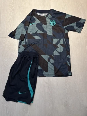Barcelona träningsset Nike svart/turkos - Snyggt träningsset från Nike med FC Barcelona-logga. Tröjan har ett geometriskt mönster i svart och turkos, rund hals och korta ärmar. Matchande svarta shorts med turkosa detaljer och Nike-logga. Perfekt för fotbollsträning eller gymmet.