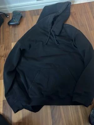 Svart hoodie med magficka - Säljer en klassisk svart hoodie med huva och dragsnören. Hoodien har en stor magficka framtill och ribbade muddar vid ärmslut och nederkant. Perfekt för en avslappnad och stilren look. Materialet känns mjukt och bekvämt.