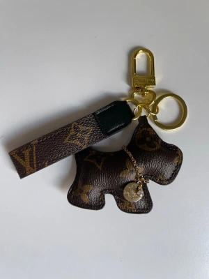 Vuitton nyckelring med hund - Snygg nyckelring från  Vuitton i brunt läder med klassiskt monogrammönster. Hunden är dekorerad med en liten guldfärgad kedja och bricka. Guldiga metalldetaljer och karbinhake. Perfekt som accessoar till väskan eller nycklarna.