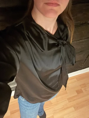 Svart satinsblus med knytband - Stilren svart satinsblus från Gina Tricot med lång ärm  med knappar. Blusen har ett snyggt knytband vid halsen som ger en elegant touch. Perfekt att styla med jeans eller kjol för en trendig look.