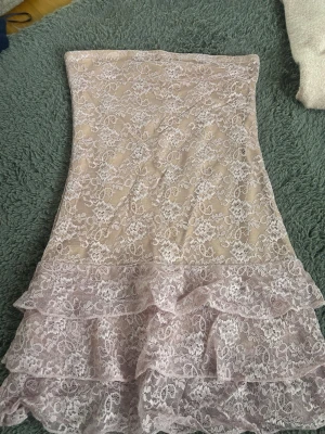 Ljus spetsklänning med volanger - Söt kort klänning i ljus färg med vacker spets och volangdetaljer nertill. Axelbandslös modell som ger en elegant och feminin look. Perfekt för dig som gillar romantisk stil.