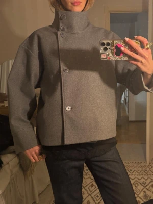 Grå boxig jacka från Gina Tricot - Säljer en grå jacka från Gina Tricot med hög krage och asymmetrisk knäppning framtill. Jackan har en boxig, oversized passform och är tillverkad i ett tjockare tyg som ger en stilren look. Perfekt för dig som gillar minimalistisk och modern stil.