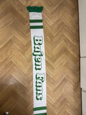 Vit och grön Bajen Fans halsduk - Säljer en vit halsduk med gröna detaljer och texten 'Bajen Fans' i stort tryck. Halsduken har gröna ränder och fransar i ena änden. Perfekt accessoar för dig som vill visa ditt stöd för Bajen på match eller i vardagen.