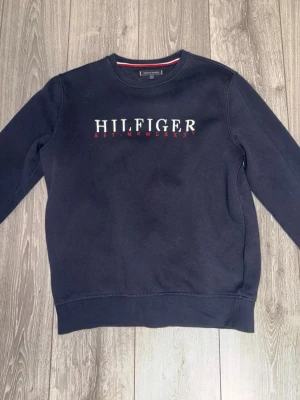 Mörkblå sweatshirt Tommy Hilfiger - Snygg mörkblå sweatshirt från Tommy Hilfiger med broderad logga och text på bröstet. Tröjan har rund halsringning och klassisk passform. Perfekt för en avslappnad och stilren look.