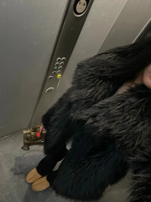 Svart fluffig pälsjacka - Säljer en svart, fluffig pälsjacka med bred krage. Jackan har en lyxig look och är perfekt för kalla dagar. Den är midjelång och har en klassisk design som passar till många olika outfits.