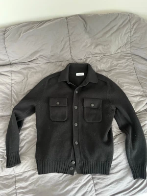 Svart cardiganjacka från Gran Sasso - Stilren svart cardiganjacka från Gran Sasso i mjukt stickat material. Jackan har två bröstfickor med knappar, ribbade muddar och knäppning framtill. Perfekt för lager på lager och en clean look. Använd några enstaka gånger så i princip ny