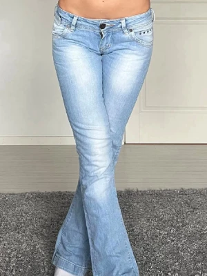 Ljusblå bootcut jeans med detaljer - Säljer ett par ljusblå bootcut jeans med slitningar och snygga detaljer vid fickorna. Jeansen har låg midja och klassisk femficksmodell. Tillverkade i mjuk denim med stretch för skön passform. Perfekta för en avslappnad och trendig look.
