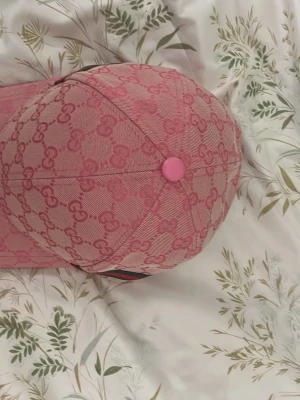 Rosa Gucci keps med monogram - Snygg rosa keps från Gucci med klassiskt GG-monogram över hela kepsen. Kepsen har böjd skärm, knapp på toppen och detaljer i grönt och rött på sidan. Tillverkad i canvasmaterial med exklusiv känsla. Perfekt för dig som vill sticka ut med en trendig accessoar.