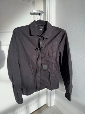 Svart overshirt från C.P. Company - Stilren svart overshirt från C.P. Company med klassisk krage, dragkedja framtill och en bröstficka med logotypdetalj. Snygg och enkel att matcha till olika outfits.