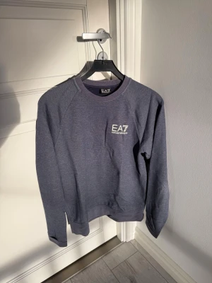 Mörkgrå sweatshirt från Armani EA7 - Snygg mörkgrå sweatshirt från EA7 Emporio Armani med rund halsringning och logga tryckt på bröstet. Tröjan har en avslappnad passform och ribbade muddar vid ärmslut och nederkant. Perfekt för en casual och sportig stil.