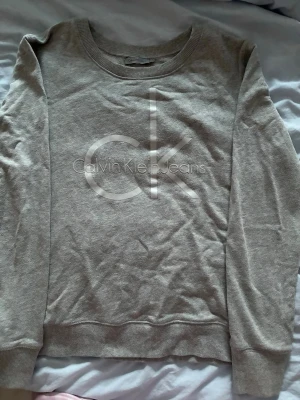 Grå sweatshirt från Calvin Klein Jeans - Säljer en stilren grå sweatshirt från Calvin Klein Jeans med stort CK-tryck framtill. Tröjan har rund halsringning, långa ärmar och ribbade muddar. Storlek L men skulle säga att den sitter som S🩷
