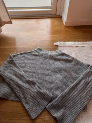 Grå ribbstickad tröja med vida ärmar - Mysig grå stickad tröja med ribbad struktur och vida ärmar. Tröjan har en avslappnad passform och hög hals, perfekt för lager på lager. Materialet känns mjukt och stretchigt, vilket gör den skön att bära under kyliga dagar.