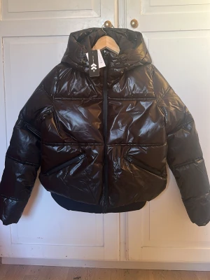 HELT NY Svart glansig pufferjacka med huva från lager  - Säljer en svart, glansig pufferjacka från lager157 med stor huva och dragkedja framtill. Jackan har två snedställda fickor och bred resår i nederkant, även en ficka på vänster ”handled” Perfekt för kalla vinterdagar och riktigt snygg till streetwear-stilen. Storlek S nypris 500 kr