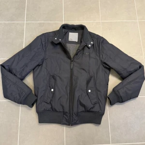 Svart vindjacka från J.Lindeberg - Stilren svart vindjacka från J.Lindeberg med ribbade muddar, ståkrage med tryckknappar och dragkedja framtill. Två snedställda fickor med knappstängning och diskret logga på bröstet. Perfekt för blåsiga dagar och enkel att matcha.