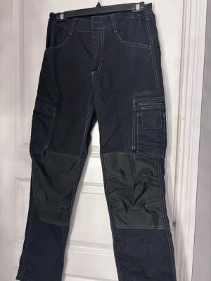 Svarta arbetarbyxor  - Svarta arbetsbyxor med vita kontrastsömmar och flera praktiska fickor på sidorna. Byxorna har förstärkta partier över knäna. Perfekta arbetsbyxor när du vill fixa till hemmet