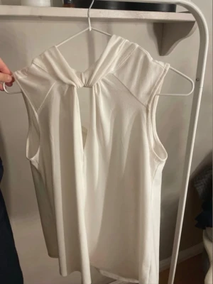 Vit ärmlös topp med knyt i halsen - Snygg vit ärmlös topp med knyt i halsen och liten öppning framtill. Toppen har en mjuk och följsam känsla, perfekt för att skapa en clean och stilren look. Passar till både jeans och kjol för en trendig outfit.